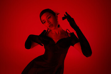 expressive asian woman with brunette hair, in black long gloves and stylish cocktail dress posing on vibrant background with red lighting effect, trendy spring, youth culture concept
