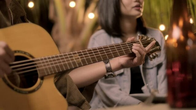 Closeup hand, A man playing acoustic guitar and sing song to girlfriend under warm lighting at night. Happy young Asian girl having fun to music and drinking together outdoor party celebrating
