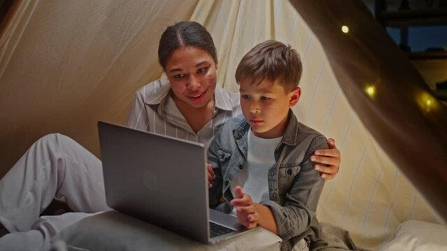 Loving mother and cute son have using laptop, browsing intenet sites, sitting inside a tent at home