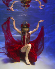 beautiful fashion girl underwater in a red fantastic dress and a designer mask is dancing under the water