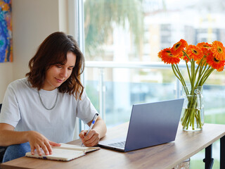 Beautiful Latin young woman in casual clothing using laptop and smiling while working indoors. writing down notes holding training for students to be executives at laptop desktop table