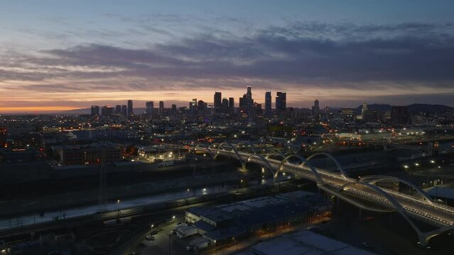 Evening cityscape shot with illuminated modern road bridge with concrete arches and silhouettes of downtown high rise buildings in background. Los Angeles, California, USA