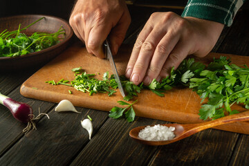 Sliced fragrant parsley for a diet salad for lunch. The hands of the cook with a knife cut fresh parsley on the kitchen board. A diet of fresh vegetables