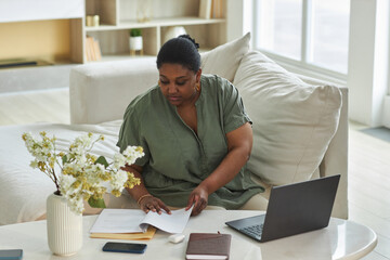 African American woman examining documents at table while working online on laptop at home