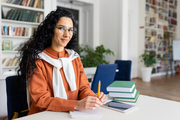 Portrait of young beautiful Latin American teacher, woman in glasses smiling and looking at camera, teaching students and pupils sitting in classroom among books.