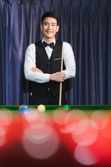 Smiling Asian snooker player arms crossed holding snooker stick There is a red snooker ball in the foreground.