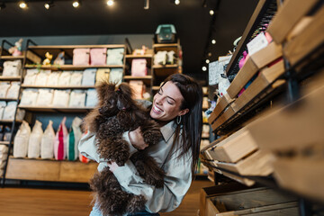 Beautiful young woman enjoying in modern pet shop together with her adorable brown toy poodle.
