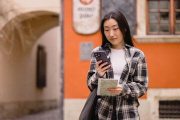 Portrait of a beautiful brunette Korean woman holding a map and comparing it with smartphone navigation on the streets of the old city. Asian tourist woman traveling in Europe.