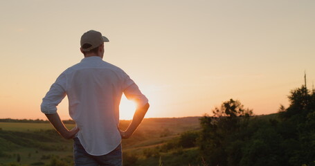 A confident young farmer stands in front of a picturesque countryside as the sun sets. View from the back