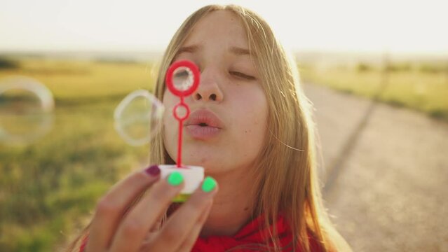 Portrait of teenager girl blowing soap bubbles on nature at sunset and smiling. Outdoors activities in summer, enjoying vacations, holidays, recreation. Positive emotions, happy childhood concept.