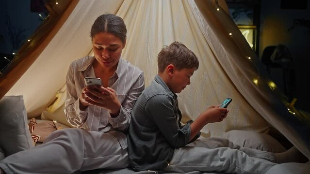 Cute child boy plays online video games while his mom checks social media content insite a game tent