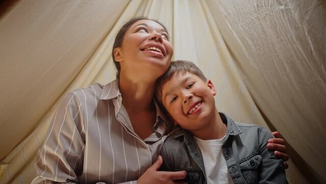 Loving mother and son sitting inside a play tent, have online video call, looking toward the camera