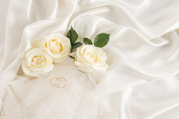 Two gold wedding rings on a beige satin background with live tea roses and part of the bride's...