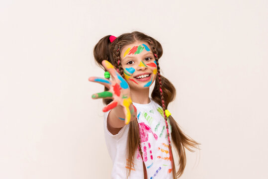 A little schoolgirl with painted hands and face with multicolored paints. A girl stained with paints. The concept of children's creativity. Funny child draws with his hands.