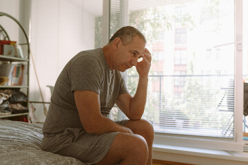 Middle-aged man in bedroom sitting on bed - obrazy, fototapety, plakaty