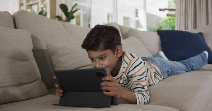 Happy biracial boy lying on sofa and using tablet at home, slow motion
