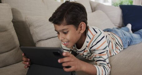 Happy biracial boy lying on sofa and using tablet at home, slow motion - Powered by Adobe