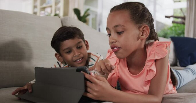 Happy biracial brother and sister lying on sofa and using tablet at home, slow motion