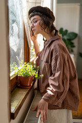 Lonely introverted hipster girl looking out window while spending time at home. Pensive young woman...