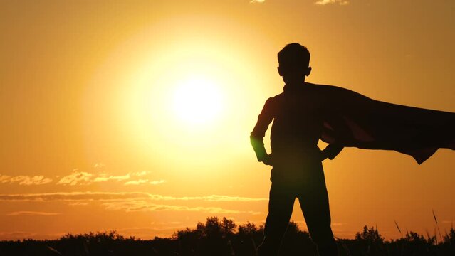 The silhouette of a superhero against the background of the sun during sunset. A kid in a superhero cloak is standing on top of a mountain, his cloak is blowing in the wind. 