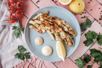 fried squid, fish pieces with white sauce, lemon and garlic. on a pink tiles table, fresh ingredients scattered around