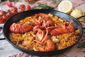 spanish seafood paella with lobster and squid, traditional dish with rice in a hot pot, surrounded by fresh ingredients on a pink background table, close up