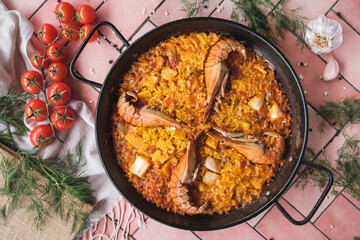 spanish seafood paella with prawns and squid pieces, traditional dish with rice in a hot pot, surrounded by fresh ingredients on a pink background table, top view