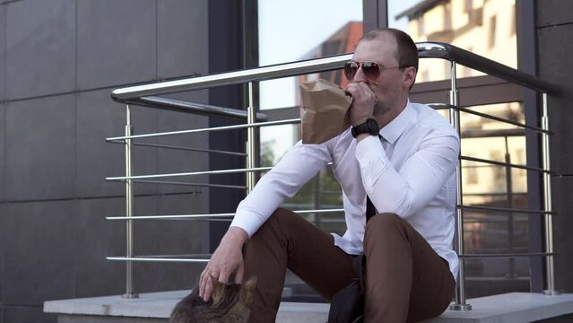 Upset man tries to calm down by breathing into a paper bag. A businessman suffering from panic attacks on the street sits near the office and pets a cat.