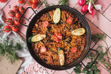 spanish paella with vegetables, zucchini, tomatoes, asparagus, traditional dish with rice in a hot pot, surrounded by fresh ingredients on a pink background table, top view