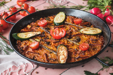 spanish paella with vegetables, zucchini, tomatoes, asparagus, traditional dish with rice in a hot pot, surrounded by fresh ingredients on a pink background table