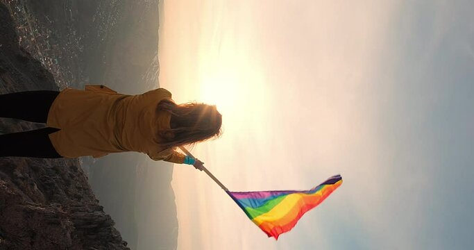 Vertical shot of a woman waving the rainbow flag standing on the top of the mountain towards the sun. Concept of courage, freedom and human rights. Back view