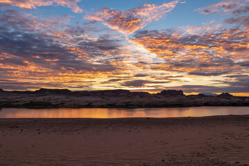 Fototapeta na wymiar Panoramic sunset view seen from Lone Rock Beach in Wahweap Bay in Lake Powell in Glen Canyon Recreation Area, Page, Utah, USA. Sand beach on wild campground. Red orange sky at sundown, warm atmosphere