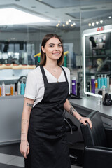 Obraz premium professional headshots, tattooed beauty worker in apron looking at camera in beauty salon, hairdressing chair, hair stylist, hair coloring, salon job, beauty salon work, hair trends