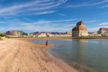 Rear view of woman walking at lakeshore looking at solitary rock formation Lone Rock in Wahweap Bay...