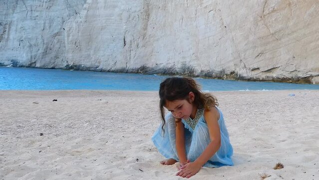 Little girl in summer blue dress playing with sand in a beach with aquamarine sea behind. High quality FullHD footage