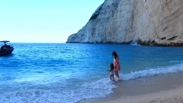 Little girl with her mother playing in water and looking on waves on a beach of aquamarine sea. High quality FullHD footage