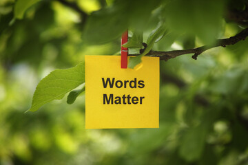 A yellow paper note with the phrase Words Matter on it attached to a tree branch with a clothes pin