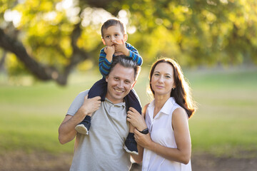 Cute white smiling family in the park - little son on the shoulders of his father and his wife standing by
