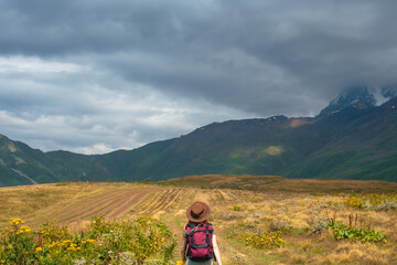 Naklejka premium Woman in wide sun hat and backpack, tourist on hiking route to Koruldi Lakes, Svaneti region, Mestia Georgia. The concept of travel and active recreation. Summer day. Rear view