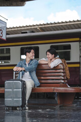 A lovely young Asian tourist couple with their suitcases enjoys talking on a bench on the platform before boarding the train together. Honeymoon trip, Summer vacation, Holidays