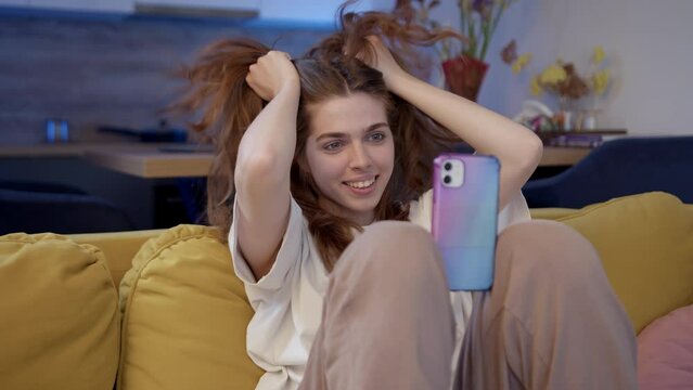 Funny beautiful girl with curly hair makes selfie while sitting on yellow sofa at home. young woman in ponytails makes ponytails and grimaces at phone camera. Video chat with girlfriend. Generation Z