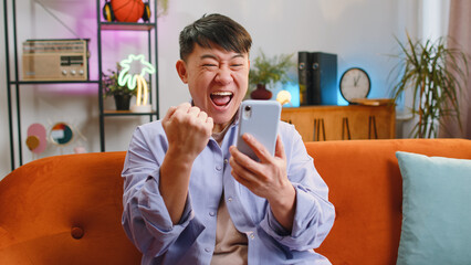 Happy asian man use mobile smartphone typing browsing say Wow yes found out great big win good news celebrate lottery jackpot doing winner gesture. Chinese adult guy at home in room sitting on sofa - Powered by Adobe
