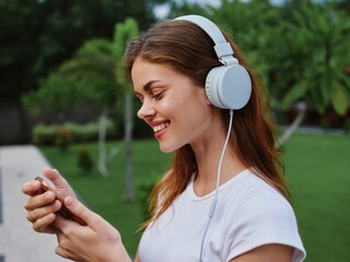 Woman wearing headphones listening to music through her phone smiling against green palm trees on a summer trip