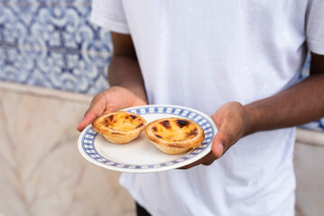 Obraz na płótnie Canvas Plate with Pastel de Nata, Portugal's traditional sweet dessert, egg custard tart pastry, in hands of young African man in front of a wall with azulejo tiles in Lisbon