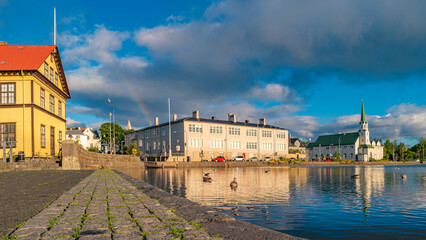 Historical and touristic downtown in Reykjavik at sunset and rainbow in Iceland. Cityscape at golden hour and blue sky at inner lake around Tjornin city park in the downtown. - obrazy, fototapety, plakaty