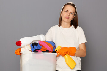 Sad upset ugappy woman wearing whote t-shirt with laundry bowl and detergent isolated over gray...