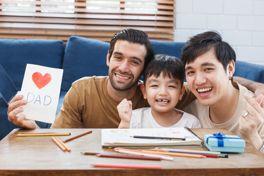 Happy cheerful LGBTQ+ lovely gay couple with their adopted child enjoy living together in living room and doing some leisure activity. Modern family and relationship lifestyle in LGBT people.