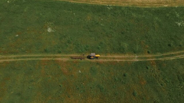Tractor with hay tedder driving by rural road between meadows after preparing fodder for cows. Work in agronomic farm for making business and production organic eco bio animal feed