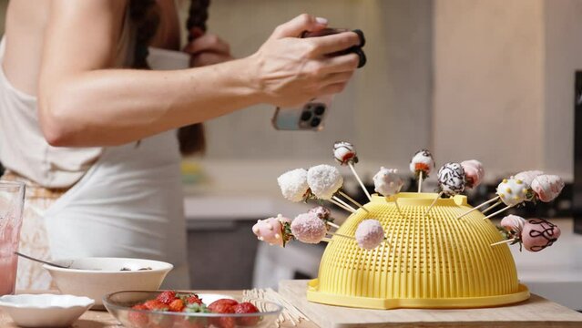 The woman has prepared strawberries dipped in chocolate and decorated with sugar sprinkles. Now she is taking a photo of them on her phone.