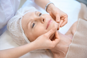 Fototapeta na wymiar Top view of mature woman in cosmetic cap getting face massage